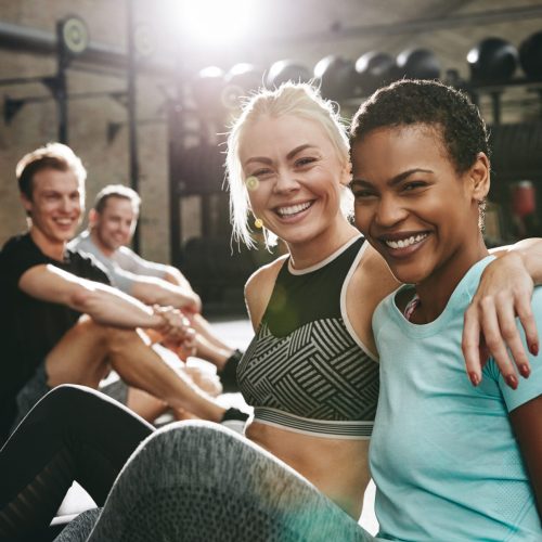 Two fit young women in sportswear laughing while sitting arm in arm together on the floor of a gym after working out