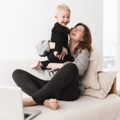 Young pretty cheerful woman sitting on sofa with her little laughing son happily playing together. Mom and baby boy joyfully spending time with laptop in cozy living room at home